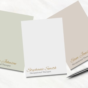 Op de afbeelding: Drie gepersonaliseerde notitieblokken in tinten groen, wit en beige, elk met een naam en de titel "Occupational Therapist" in goudkleurig schrift. Een zilveren pen ligt op het oppervlak.