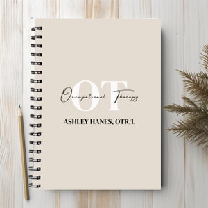 May include: A spiral-bound notebook with a beige cover. The cover features the text "OT Occupational Therapy" in a mix of serif and script fonts, along with the name "ASHLEY HANES, OTR/L". A pencil rests on the wooden surface.