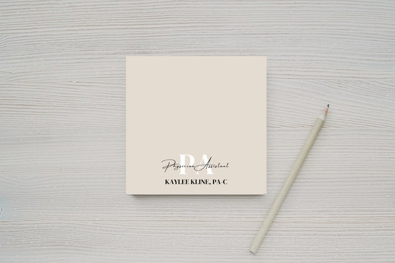 May include: A beige notepad with the text "PA Physician Assistant KAYLEE KLINE, PA-C" printed on it. A pencil is lying on a white wooden surface next to the notepad.