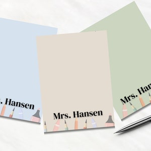 Peut inclure: Trois blocs-notes vierges avec un fond bleu clair, beige et vert. Chaque bloc-notes a une rangée de crayons de couleur en bas et le texte "Mrs. Hansen" en bas.