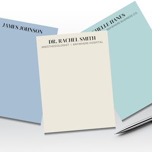 May include: Three blank notepads with different coloured backgrounds. The top notepad is light blue with the text "JAMES JOHNSON" printed on it. The middle notepad is cream coloured with the text "DR. RACHEL SMITH ANESTHESIOLOGIST | ANYWHERE HOSPITAL" printed on it. The bottom notepad is light blue with the text "CHELLE HANES ANYWHERE BUSINESS CO." printed on it.