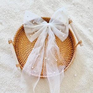 May include: A white tulle bow with pearl embellishments, displayed on a woven wicker tray.