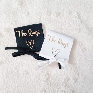 May include: Two ring bearer pillow boxes, one black and one white, with gold lettering that says "The Rings" and a gold heart. The boxes are on a white fluffy surface.