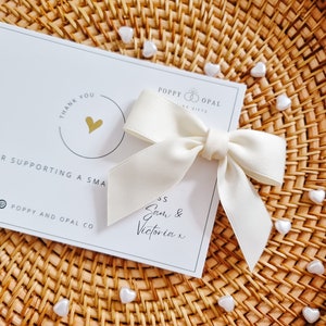 May include: A white card with the text "Thank You" and "Poppy and Opal Co."  A white satin bow is tied on top of the card. The card is sitting on a woven wicker surface with white heart-shaped beads scattered around it.