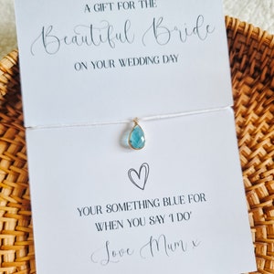 Puede incluir: Una tarjeta blanca con el texto "Un regalo para la hermosa novia en el día de tu boda." Un colgante de piedra azul cuelga de una cuerda blanca. La tarjeta también dice "Tu algo azul para cuando digas 'Sí, quiero' Con amor Mamá x"