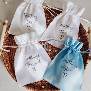 May include: Four small white drawstring bags with the words "Something Old", "Something New", "Something Borrowed", and "Something Blue" printed on them. The bags are sitting in a woven basket.