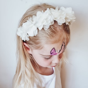 May include: A young girl wearing a white flower headband. The headband is made of fabric flowers and is worn around the girl's head. The girl has blonde hair and is looking down.