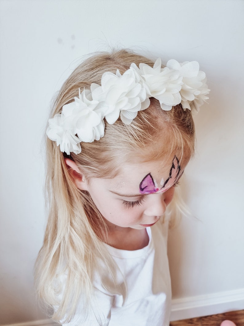 May include: A young girl wearing a white flower headband. The headband is made of fabric flowers and is worn around the girl's head. The girl has blonde hair and is looking down.