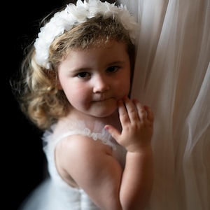 May include: A young girl wearing a white dress and a white flower crown, looking at the camera with a slight smile.