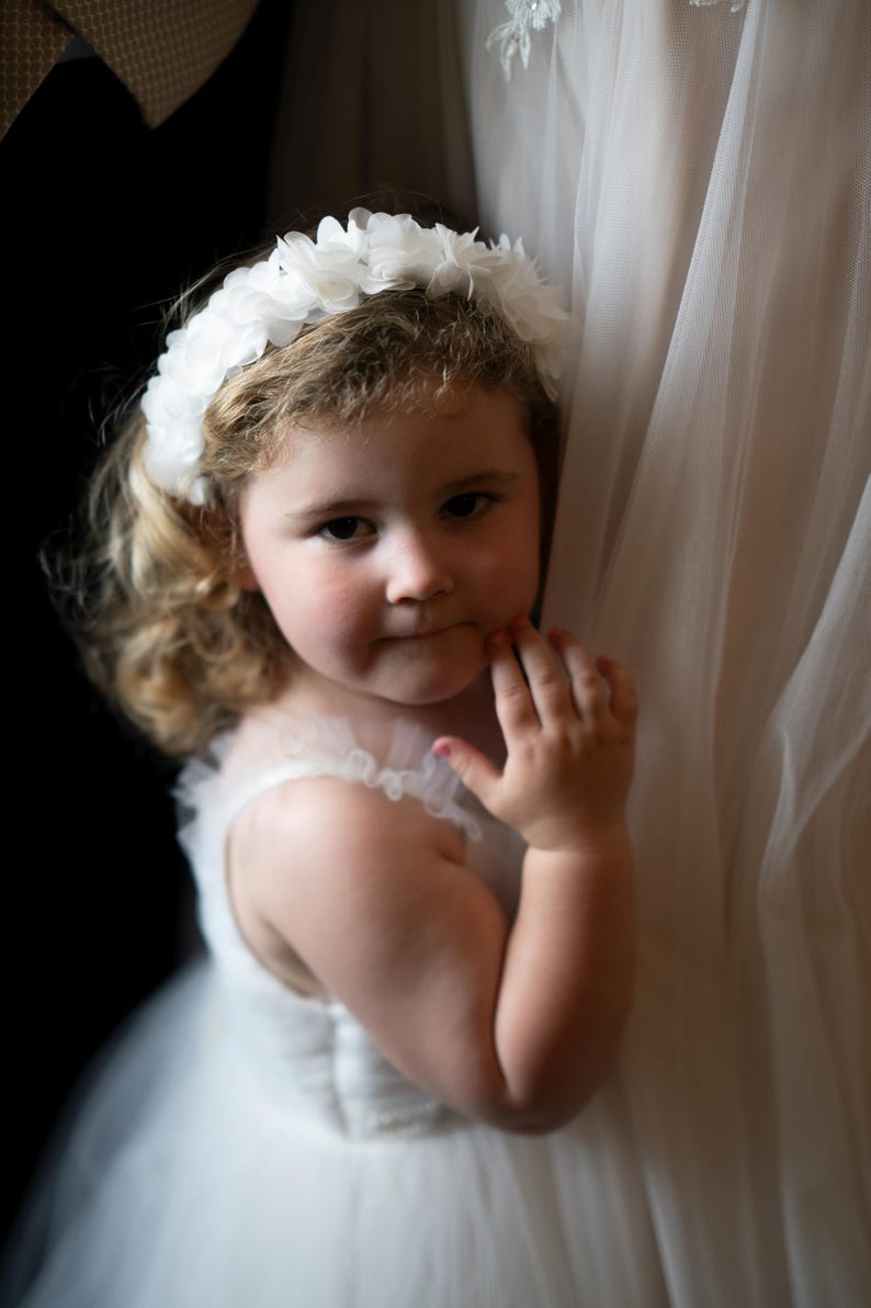 May include: A young girl wearing a white dress and a white flower crown, looking at the camera with a slight smile.