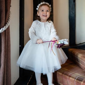 May include: A young girl in a white tulle dress with lace details and a white flower crown smiles while holding a bouquet of flowers.