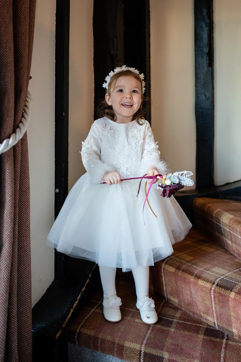 May include: A young girl in a white tulle dress with lace details and a white flower crown smiles while holding a bouquet of flowers.