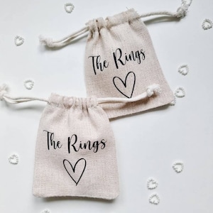 Puede incluir: Dos pequeñas bolsas de tela beige con cordón de ajuste con el texto "The Rings" y un corazón impreso en ellas. Las bolsas están sobre un fondo blanco con pequeños confeti blancos en forma de corazón.