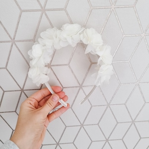 May include: A white headband with a floral design made of white fabric flowers. The headband is made of a thin, flexible material.