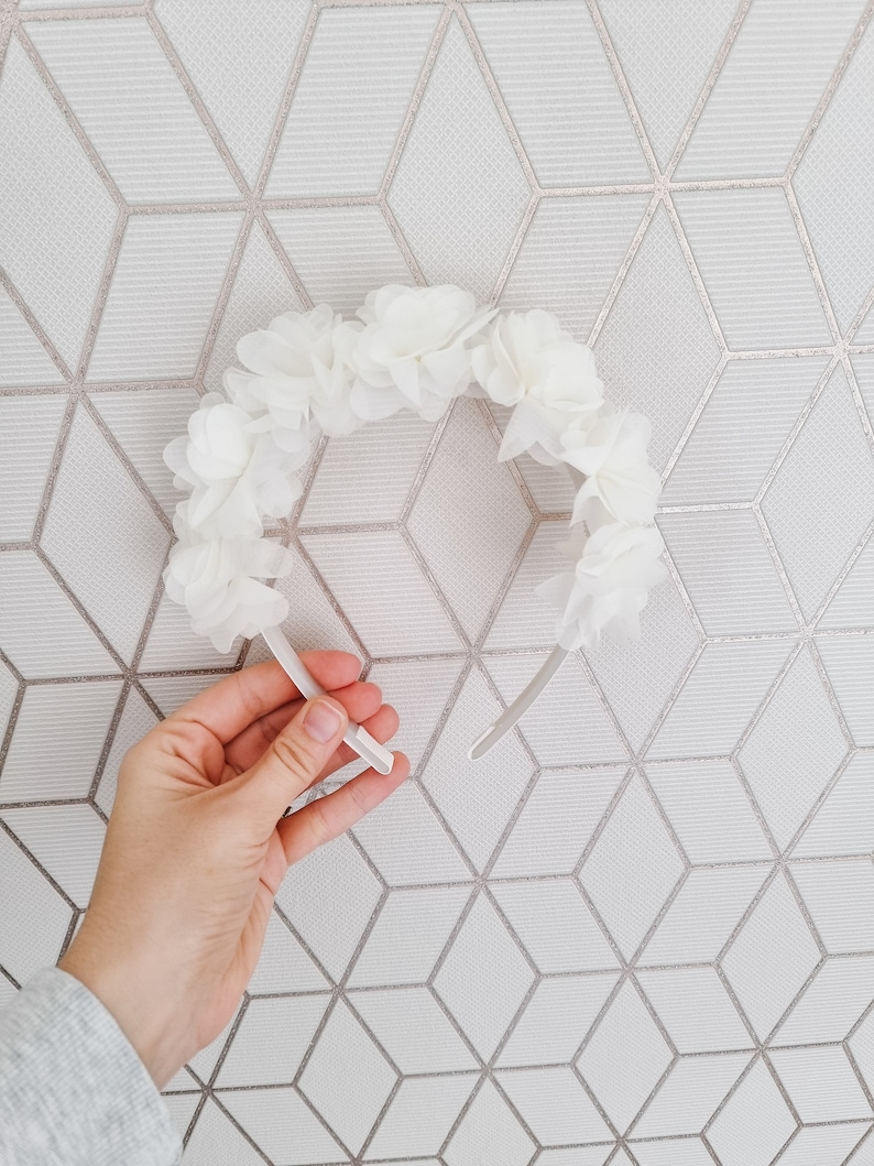 May include: A white headband with a floral design made of white fabric flowers. The headband is made of a thin, flexible material.