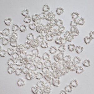 May include: A scattering of small, white heart-shaped beads on a white background. Each heart is formed from tiny, round beads, creating a textured outline. The beads are evenly spaced, giving a clean and elegant appearance, suitable for crafts.