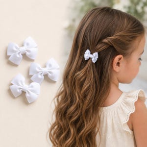 Könnte beinhalten: Ein Set aus kleinen weißen Haarschleifen aus Ripsband. Drei einzelne Schleifen sind separat abgebildet, und eine identische weiße Schleife ist an einer Partie langen, welligen braunen Haares befestigt, die seitlich mit einer Spange fixiert ist.