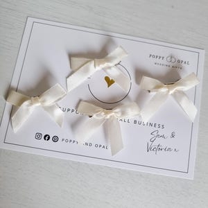 May include: Four ivory satin hair bows with gold clips. The bows are arranged on a white card with the text "Poppy and Opal Wedding Gifts" and "Small Business Sam & Victoria x".