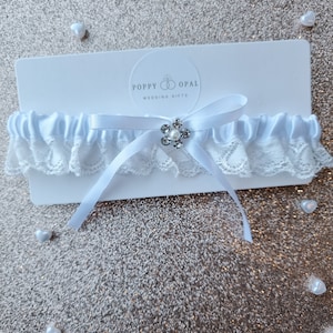 Op de afbeelding: Witte satijnen kousenband met een witte kantversiering en een zilveren strass-bloemversiering. De kousenband ligt op een witte kaart met de tekst "Poppy Opal Wedding Gifts".
