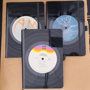 May include: Three black notebooks with vinyl record designs. The notebooks feature album art from the bands Kansas, Waylon, and Styx. Each notebook has an elastic band closure and a unique album label design.