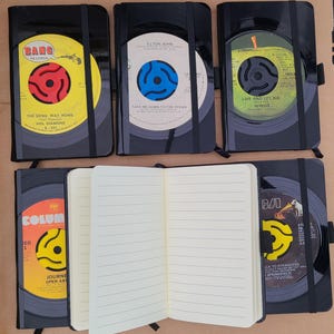 May include: Black notebooks with vinyl record designs. The notebooks feature records from artists like Elton John and Wings. One notebook is open, revealing lined pages. The covers display record labels with text and colorful graphics.