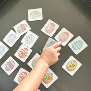 May include: A child's hand reaches for a card with a blue and white patterned egg. The card is part of a set of matching cards featuring different patterned eggs. The cards are laid out on a gray surface.