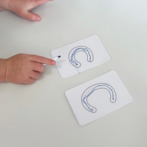 May include: Two white cards with blue lines tracing the letter "C" on them. A child's hand is tracing the letter on one of the cards with a white marker.