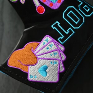 May include: Black leather wallet with colorful embroidered patches, including a hand holding a set of four aces, a slot machine, and the word "LUCK" in blue.