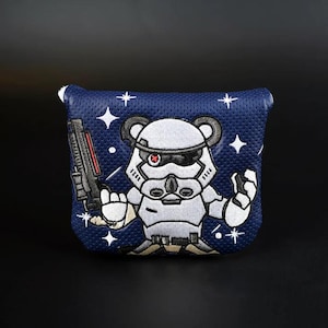 Pode incluir: Uma capa para cabeça de taco de golfe azul e branca com um personagem de desenho animado de um Stormtrooper com um olho vermelho. O personagem está segurando um blaster e tem um tema de Star Wars.