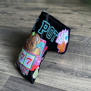 May include: Black golf club head cover with a colorful embroidered design featuring a cartoon bear, slot machine, and playing cards. The text "LUCKY POT" is embroidered on the cover.