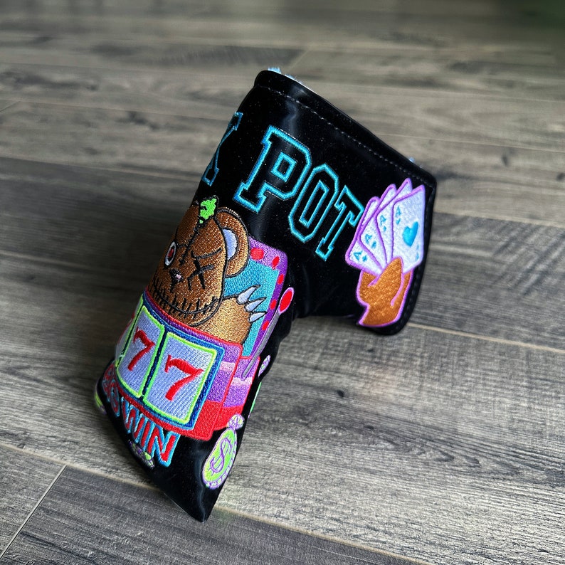 May include: Black golf club head cover with a colorful embroidered design featuring a cartoon bear, slot machine, and playing cards. The text "LUCKY POT" is embroidered on the cover.