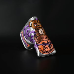 May include: A silver golf club head cover with a purple and orange design. The design features a cartoon teddy bear with a red eye, sitting on top of a stack of money. The money is tied with a purple rope.