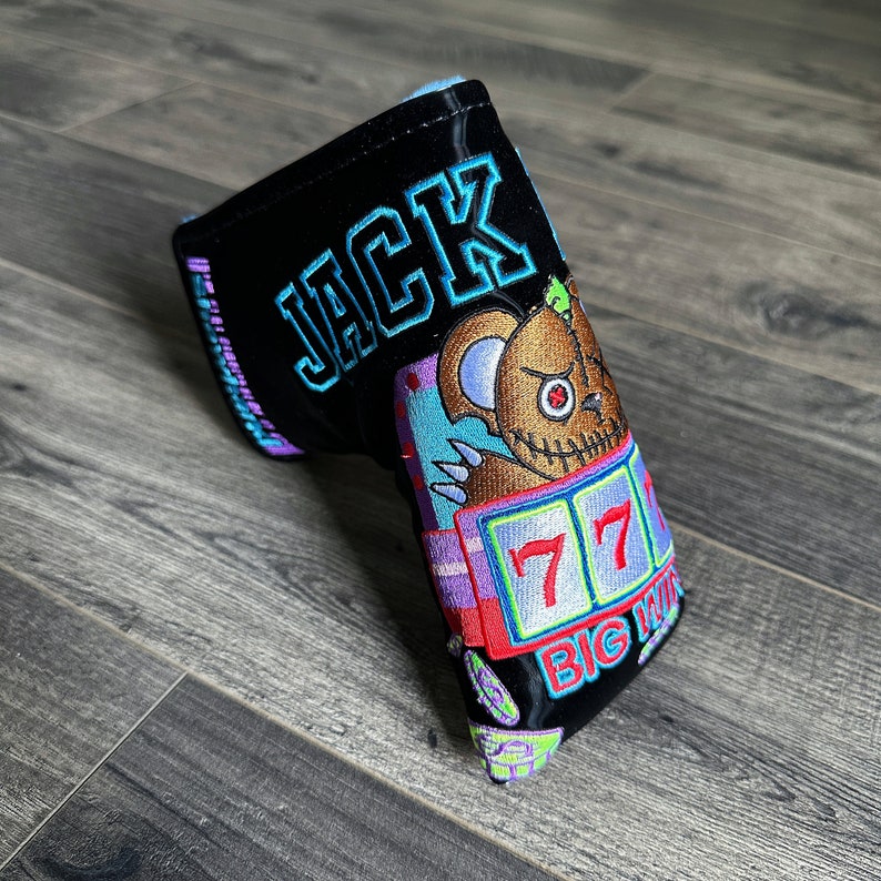 May include: Black golf club head cover with a colorful embroidered design featuring a cartoon bear, a slot machine with the number 77, and the text "BIG WIN". The cover also has the name "JACK" embroidered on it.