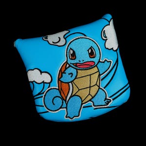 May include: A blue golf club headcover featuring an embroidered image of Squirtle, a cartoon turtle character, with a tan shell and blue body. The cover has a blue sky background with white cloud accents and black outlines.