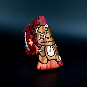 May include: A red and orange golf club head cover with a stitched teddy bear design. The bear has a stitched-up mouth, one eye, and a red cross on its chest. The text "Showhand" is on the side of the cover.