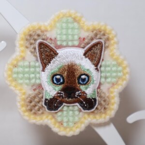 May include: Embroidered patch featuring a Siamese cat face with blue eyes, brown ears, and a white face. The cat is centered on a flower-shaped, cross-stitched background in pastel colors of yellow, green, and brown. The patch is likely for clothing or accessories.