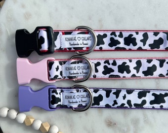 May include: Three pet collars featuring a black and white cow print pattern. Each collar has a side-release buckle in black, light pink, or light purple, a metal D-ring, and a white fabric label reading KONAKAI COLLARS Handmade in Hawaii.