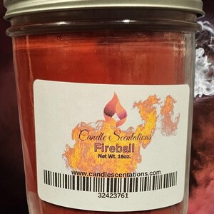 Pu&ograve; includere: Una candela rossa in un barattolo di vetro con un'etichetta bianca che recita "Candle Scentations Fireball Net Wt. 16oz. www.candlescentations.com 32423761".