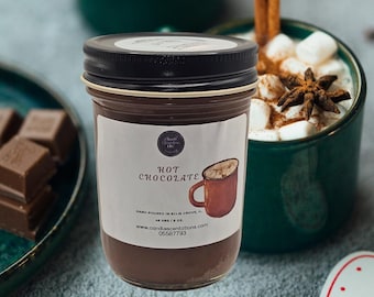 Könnte beinhalten: Ein Glasgefäß gefüllt mit einem dunkelbraunen Heißschokoladen-Mix. Das Gefäß hat einen schwarzen Deckel und ein weißes Etikett mit der Aufschrift "Hot Chocolate". Das Gefäß steht auf einem Tisch mit einer Tasse Heißschokolade und Marshmallows im Hintergrund.