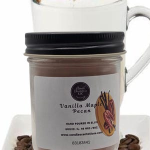 Könnte beinhalten: Eine Glas-Kerze in einem Einmachglas mit brauner Wachsgrundlage und einem weißen Etikett. Das Etikett lautet "Candie Scentations LLC", "Vanilla Maple Pecan", "Hand Poured in Ellis Grove, IL. 48 Hrs/8oz.", "www.candlescentations.com" und "83183441". Das Glas steht auf einem weißen Teller mit Kaffeebohnen.