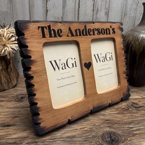 May include: A rectangular wooden double photo frame with a burnt, charred edge finish. The top is engraved with the text "The Anderson's" and a small heart icon sits between the two photo openings. Each opening contains a paper insert with the text "WaGi" and "Your Photo Goes Here".