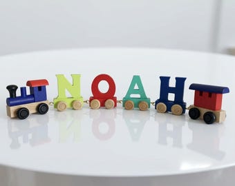 May include: A wooden toy train set featuring a blue engine and a red caboose connected to four individual letter cars that spell the name NOAH. Each letter car is painted in a different colour, including lime green, red, teal, and dark blue.