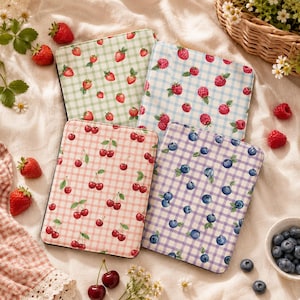 May include: Four rectangular e-reader covers featuring gingham patterns in light green, light blue, pink, and light purple. Each cover displays a repeating fruit print: strawberries on the green, raspberries on the blue, cherries on the pink, and blueberries on the purple. The covers are arranged in a grid on a white fabric surface.