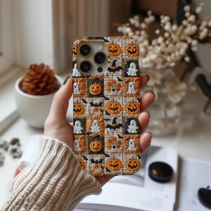 May include: A phone case with a Halloween theme, displaying a patchwork pattern of orange pumpkins, white ghosts, and black bats set against a plaid backdrop. The case is held in a hand, highlighting the autumnal design.