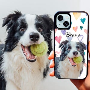 Op de afbeelding: Een zwart-witte border collie met een tennisbal in zijn bek is afgebeeld op een witte telefoonhoes met een aquarelontwerp van harten en de naam "Bruno".