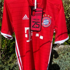 Pu&ograve; includere: Una maglia da calcio Adidas rossa con dettagli bianchi, appesa all'aperto. La maglia presenta il logo del Bayern Monaco, il logo Adidas e il nome "MULLER 25" su una custodia per telefono agganciata. Strisce bianche corrono lungo i lati.