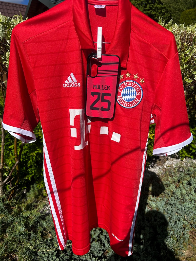 Pu&ograve; includere: Una maglia da calcio Adidas rossa con dettagli bianchi, appesa all'aperto. La maglia presenta il logo del Bayern Monaco, il logo Adidas e il nome "MULLER 25" su una custodia per telefono agganciata. Strisce bianche corrono lungo i lati.