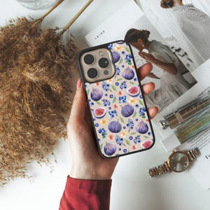 Puede incluir: Una funda de teléfono negra con un estampado de higos y flores. El diseño incluye higos enteros y cortados, arándanos y pequeñas flores en tonos morados, azules y amarillos sobre un fondo beige. La funda se sostiene en una mano.