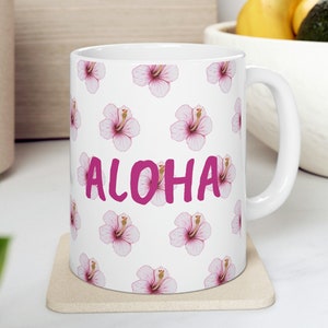 Könnte beinhalten: Eine weiße Keramiktasse mit einem Muster aus rosa Hibiskusblüten und dem Wort "ALOHA" in rosa Schrift. Die Tasse steht auf einem beigefarbenen Untersetzer.