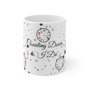 Puede incluir: Taza de cerámica blanca con un patrón de salpicaduras de colores y gráficos de reloj. El texto "Counting Down to 'I Do'" está escrito en cursiva negra.